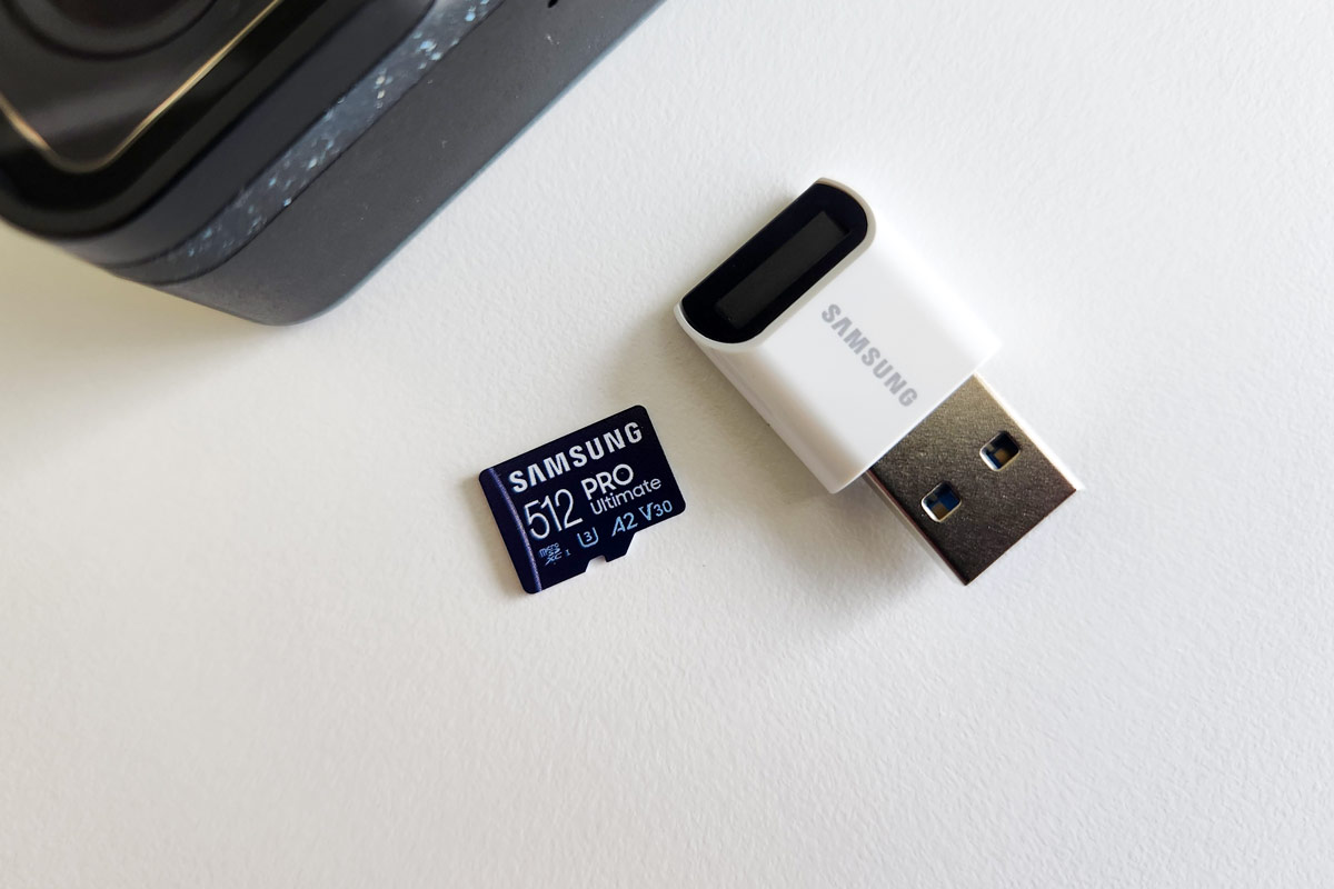 Samsung PRO Ultimate 512GB microSDXC memory card for cameras next to a microSDXC card reader and GoPro action camera on a white desk.