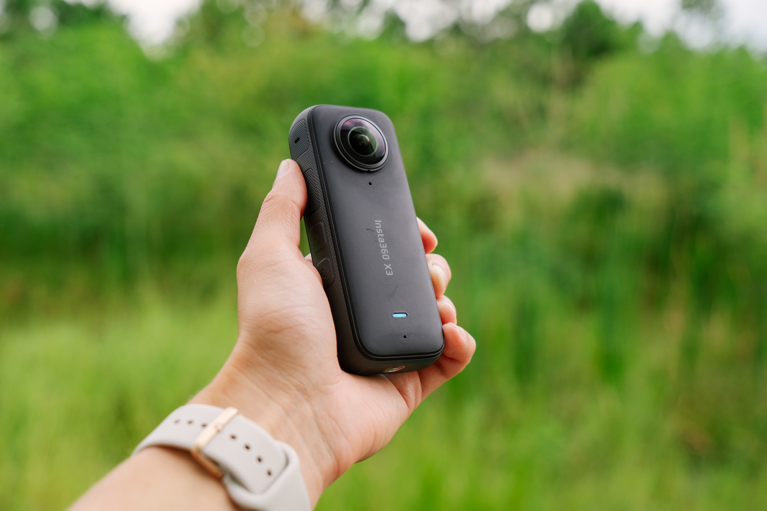 The Insta360 X3 held in a hand in front of a green forest