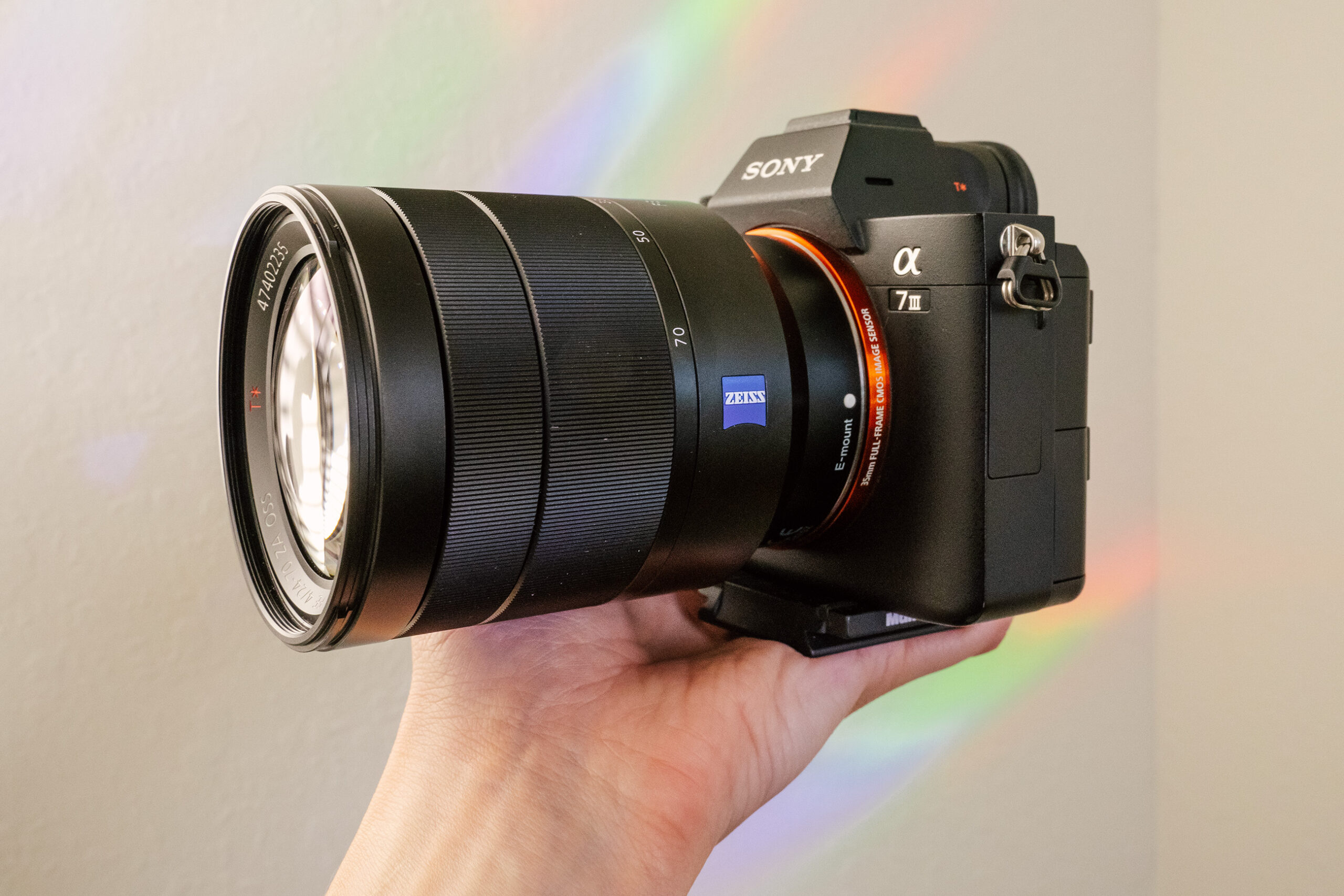 A hand holds the Sony Vario-Tessar T* FE 24-70mm f/4 Lens and Sony a7 III camera in front of a white wall with rainbows.